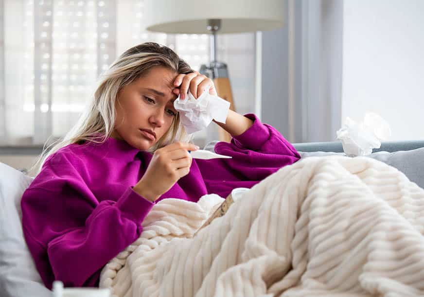 a sick woman sits on her couch under a blanket