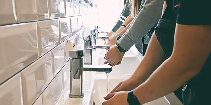 Three people washing their hands in a porcelain sink with metal faucets.