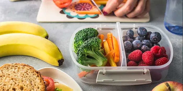 A plastic two-compartment lunch container filled with a sandwich, broccoli, carrots, and berries, next to strawberries and a cutting board where a person’s hands are slicing fruit.