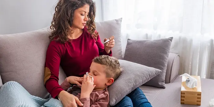 Mother caring for sick child on couch, checking temperature and helping him blow his nose during flu or COVID season.