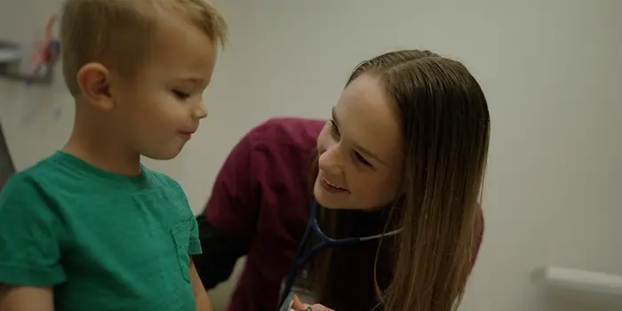 Urgent care clinician smiling at a young child while using a stethoscope during a checkup.
